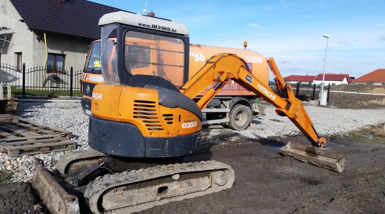Pomarańczowa mini koparka Hitachi EX30UR na ziemi, w tle pomarańczowy samochód z napisem Bucher i domy. Widoczne palety i kamienie.