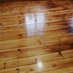 Odnowiona, lakierowana podłoga z desek sosnowych z widocznymi śladami po gwoździach, ceglana ściana w tle i grzejnik.