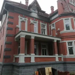 Odrestaurowana fasada budynku z czerwonej cegły z białymi oknami, widoczne pozostałości po pracach remontowych i stare drzwi leżące na ziemi.