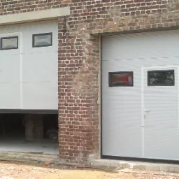 Dwie szare bramy segmentowe z małymi okienkami w czarnych ramkach, zamontowane w budynku z czerwonej cegły, jedna brama częściowo otwarta.