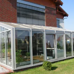 Szklana oranżeria z białymi ramami przylegająca do ceglanego domu, widok z zewnątrz na zadbany trawnik i rośliny wewnątrz.