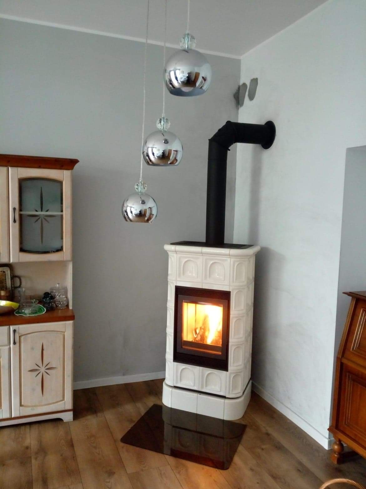 Biały, kaflowy piec wolnostojący z czarnym kominem w rogu pokoju, z palącym się ogniem, podwieszonymi srebrnymi kulami i drewnianą podłogą.