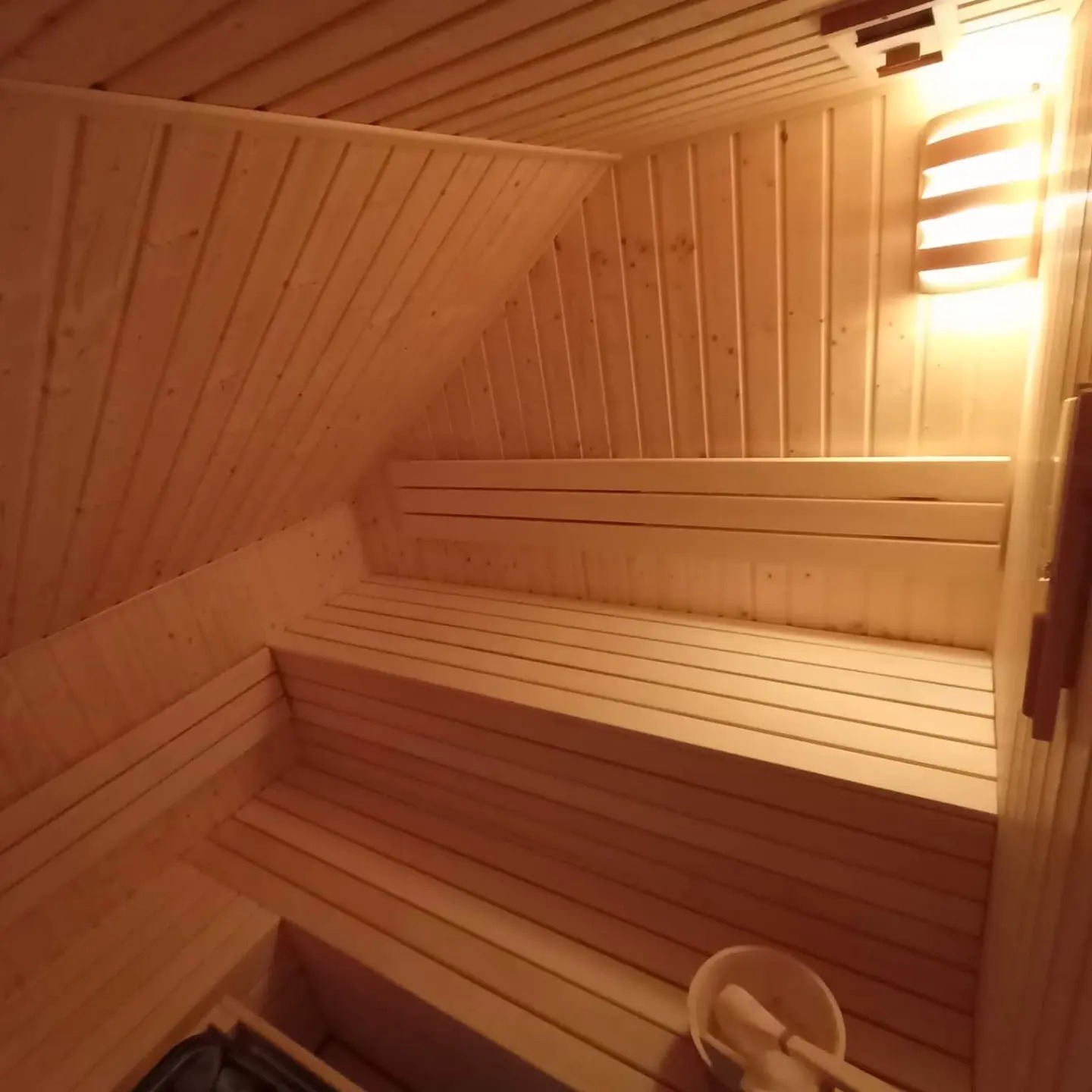 Wnętrze sauny wykończone jasnym drewnem, z widocznymi ławkami, oświetleniem i akcesoriami do polewania kamieni. W tle skośny sufit.
