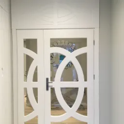 Białe dwuskrzydłowe drzwi wewnętrzne z przeszklonymi dekoracyjnymi elementami, zamontowane w jasnym pomieszczeniu z drewnianą podłogą.