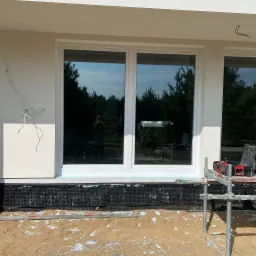 Montaż okna PCV w nowym budynku, widoczne rusztowanie, poziomica i folia fundamentowa, odbicie fotografa w szybie.