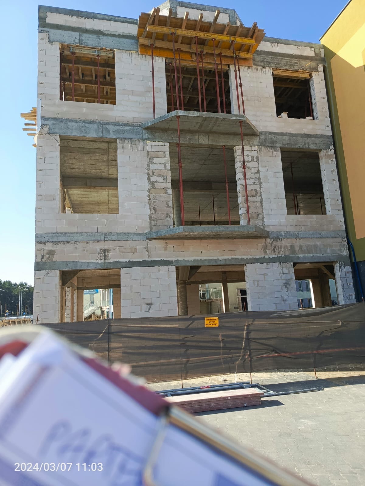 Wznoszony budynek z betonu komórkowego, widoczne otwory okienne i drzwiowe, wsporniki stropowe, częściowo zasłonięty siatką budowlaną i dokumentami w kadrze.