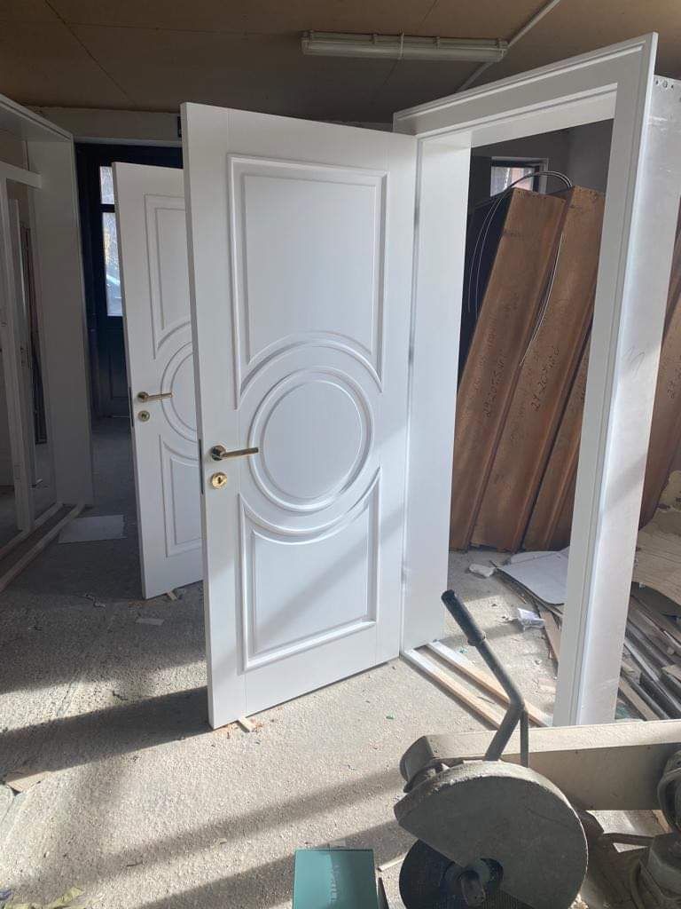 Białe drzwi wewnętrzne z okrągłym zdobieniem, uchwytem w kolorze złota, uchylone, widoczne w warsztacie stolarskim z narzędziami i materiałami.