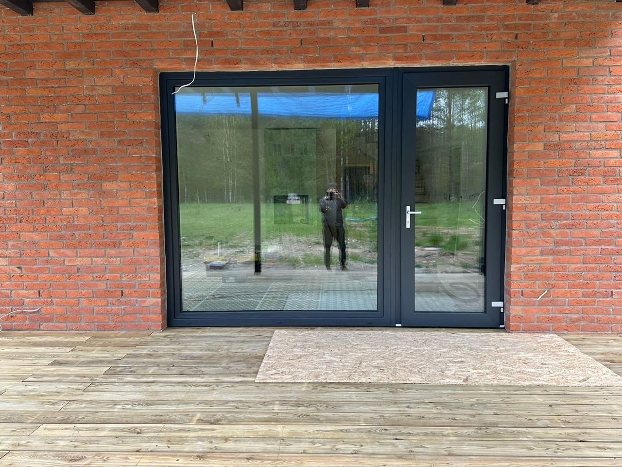 Szare przeszklone drzwi balkonowe z ramą z tworzywa sztucznego w murze z czerwonej cegły, widoczne odbicie fotografa i fragment drewnianego tarasu z płytą osb.