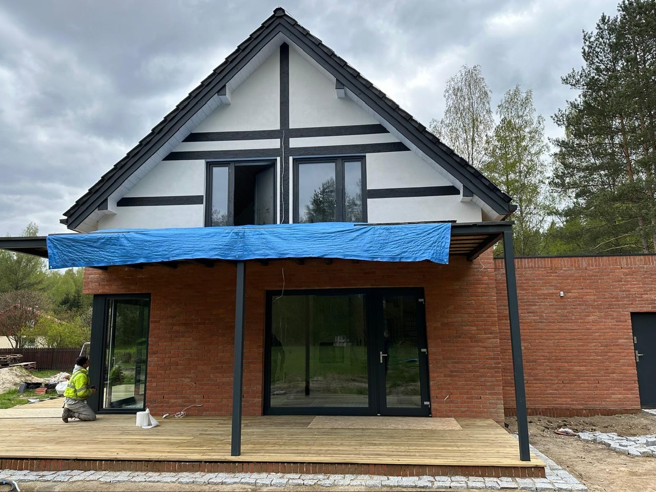 Montaż okien w nowym domu z cegły, widoczna osoba w odblaskowej kamizelce pracująca na drewnianym tarasie, fragment okna otwarty, zabezpieczony niebieską plandeką.