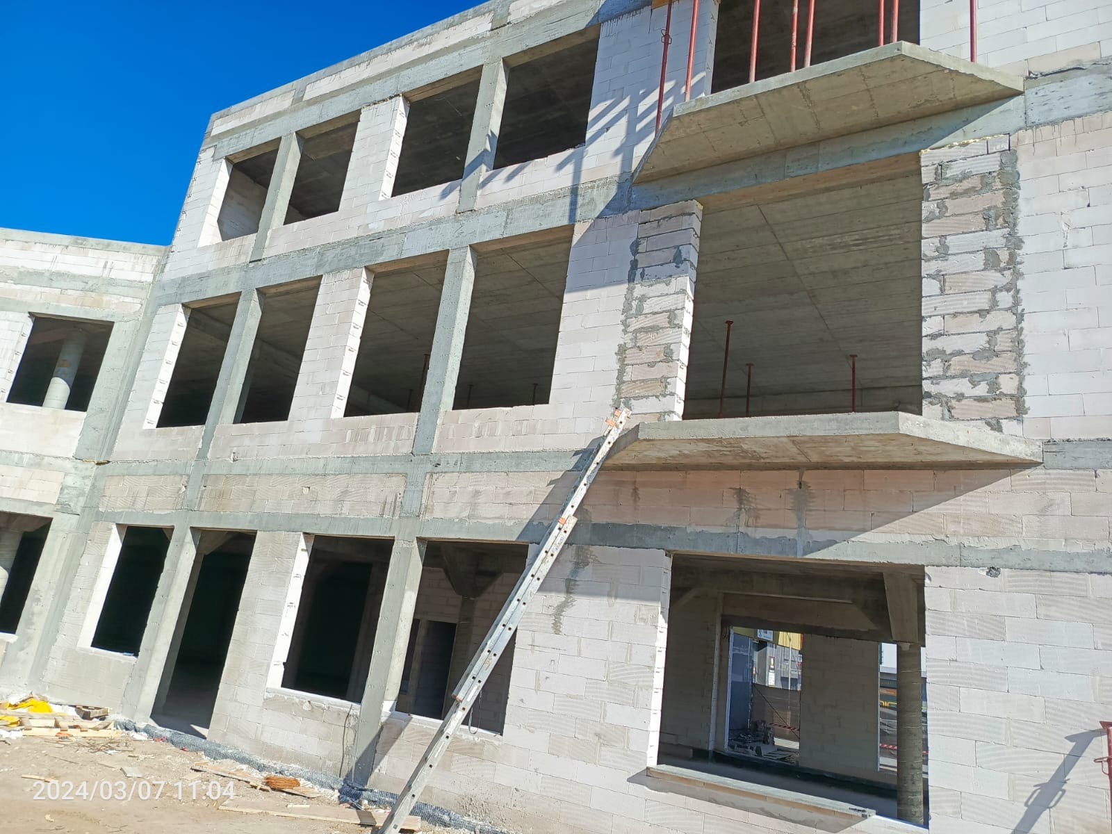 Surowy budynek wielopiętrowy w trakcie budowy, z widocznymi otworami okiennymi i balkonami, drabina oparta o ścianę, błękitne niebo w tle.