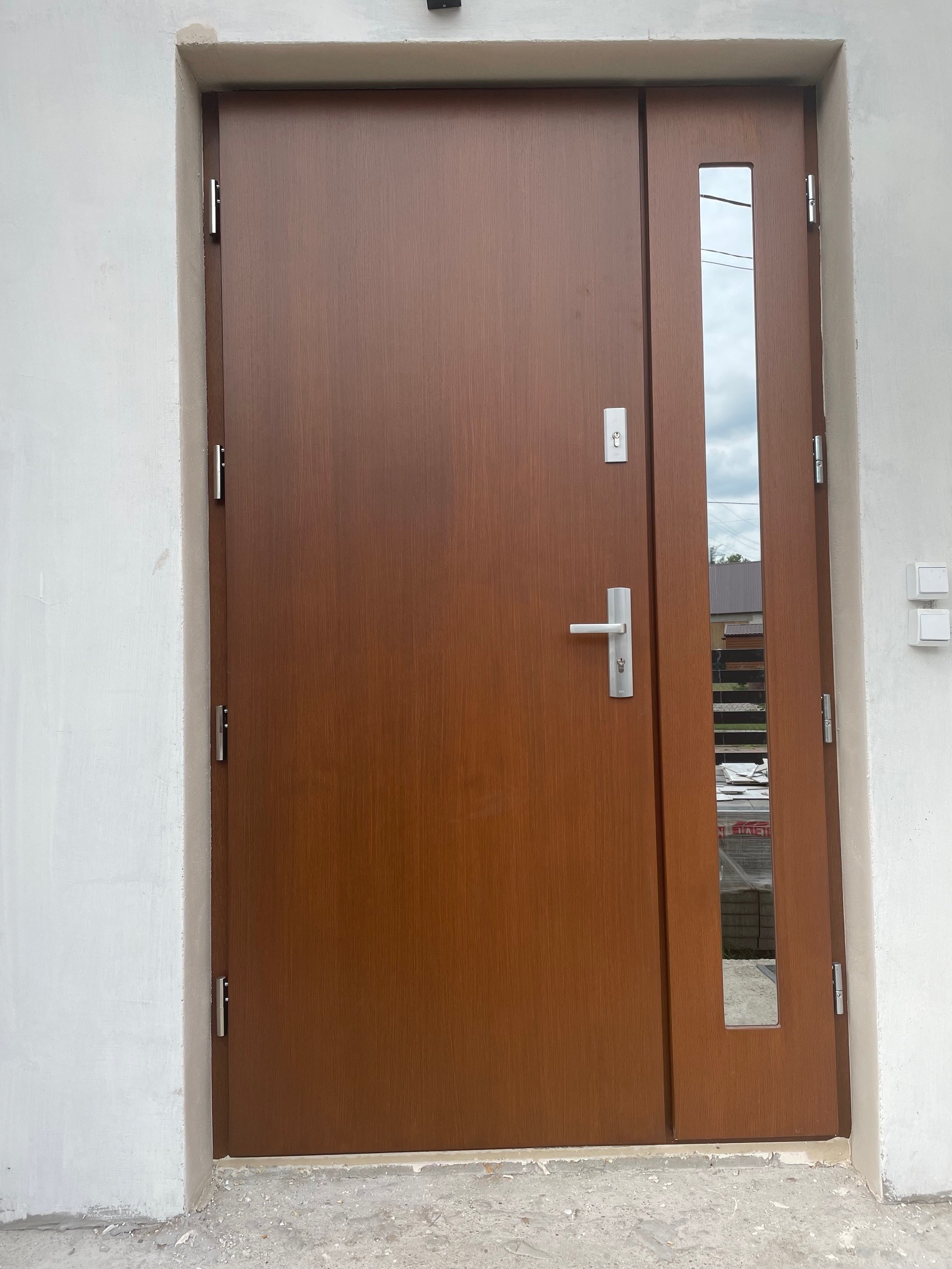 Brązowe drzwi wejściowe z długim, pionowym przeszkleniem, zamontowane w surowym, otynkowanym otworze ściennym, widoczne zawiasy i klamka.