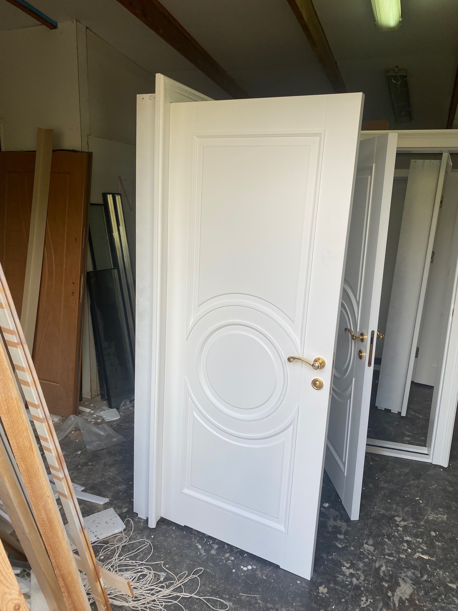 Białe, lakierowane drzwi wewnętrzne z okrągłym zdobieniem i złotą klamką, ustawione w magazynie lub warsztacie, widoczne inne drzwi i elementy drewniane.