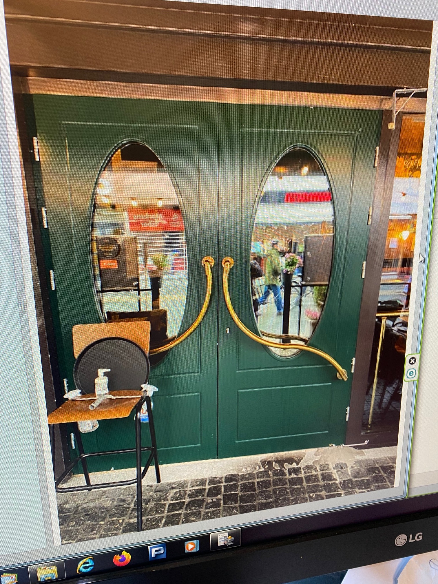 Zielone, dwuskrzydłowe drzwi wejściowe z owalnymi przeszkleniami i zakrzywionymi, złotymi uchwytami; przed drzwiami stoi stolik z płynami do dezynfekcji.