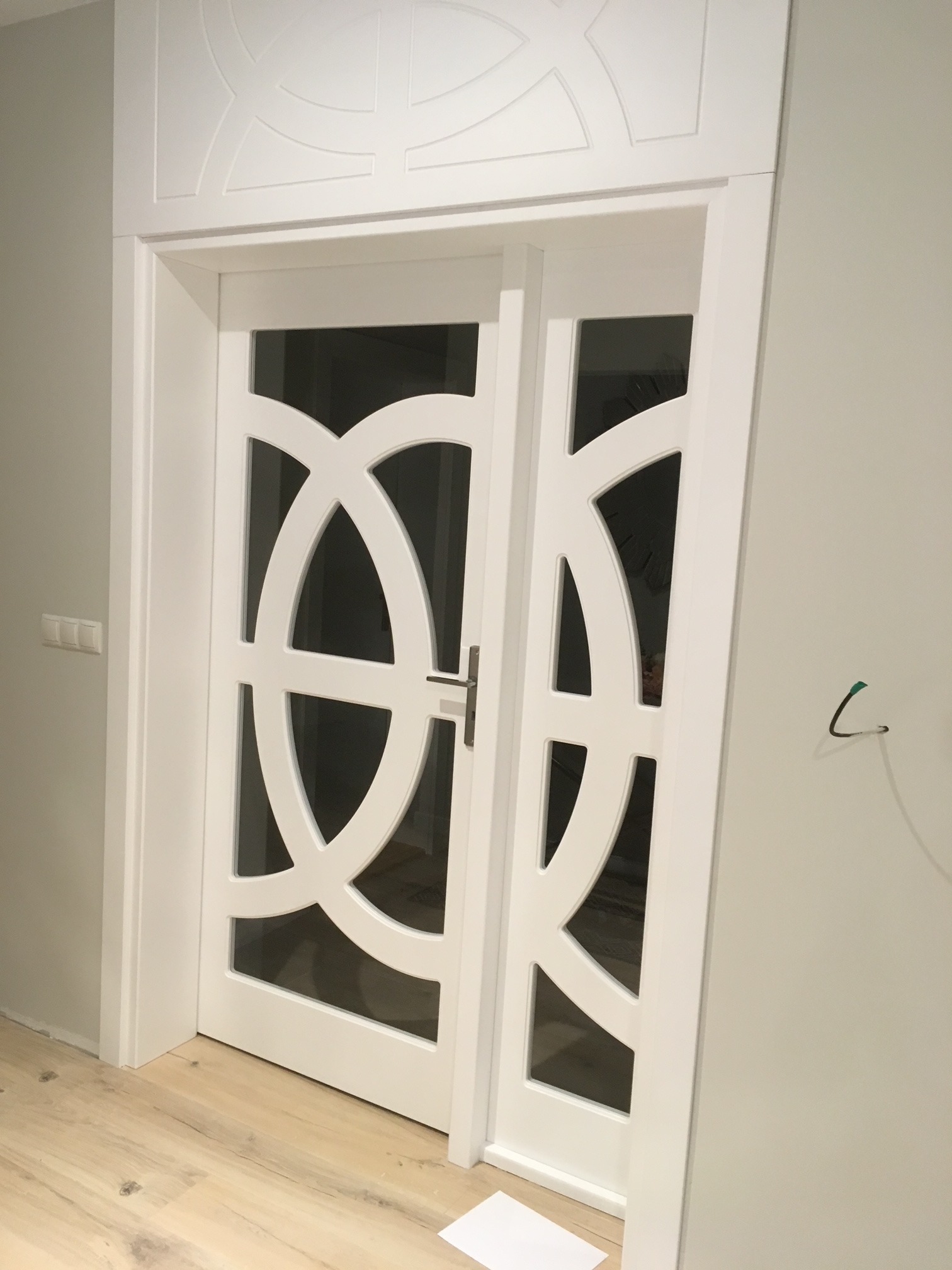 Białe drzwi wewnętrzne z przeszkleniami o geometrycznym wzorze, widoczne na tle jasnej ściany i drewnianej podłogi, z dekoracyjnym elementem nadproża.