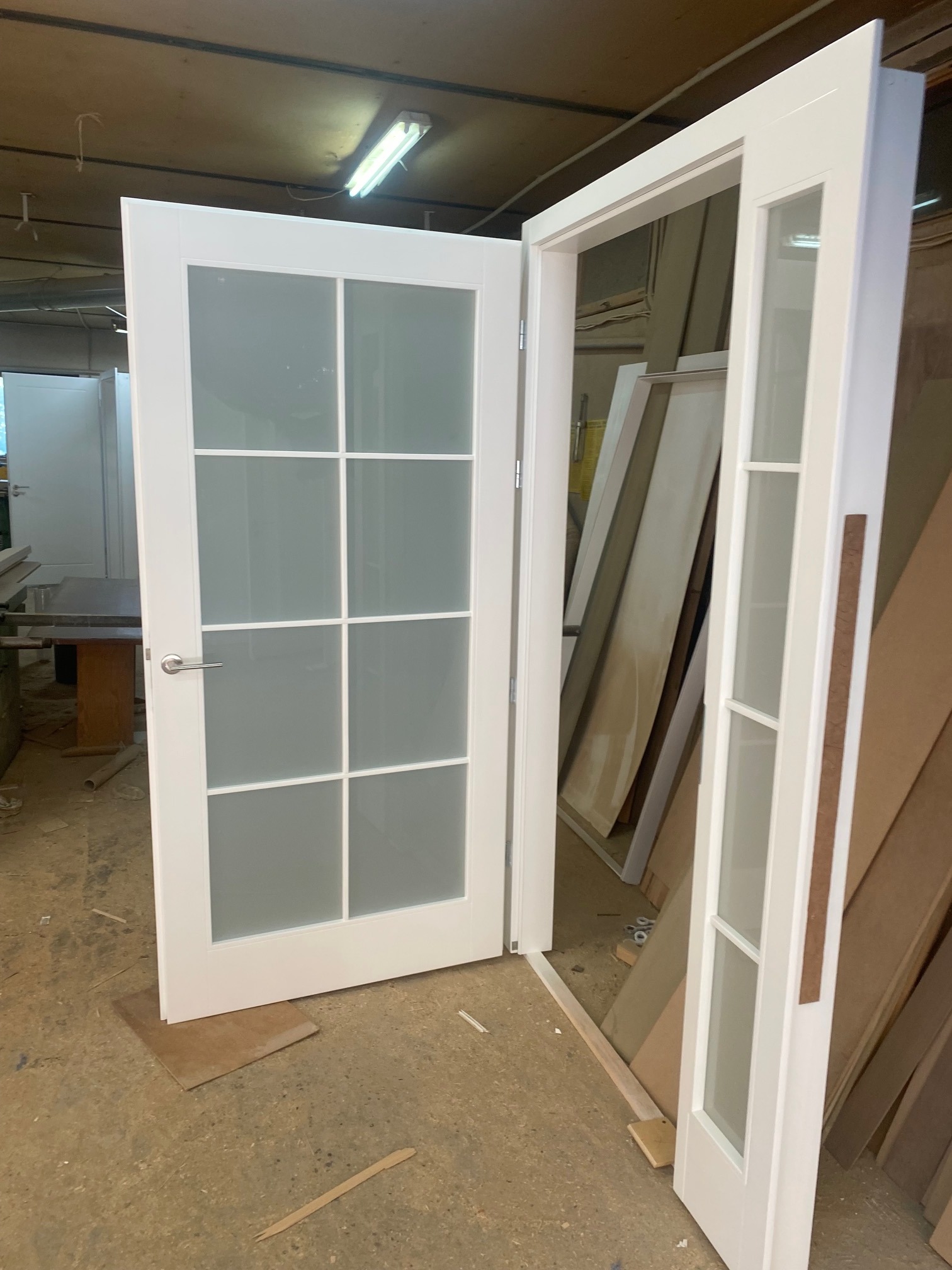 Białe, przeszklone drzwi z ośmioma szybkami, częściowo otwarte w warsztacie, widoczne płyty i materiały budowlane w tle.