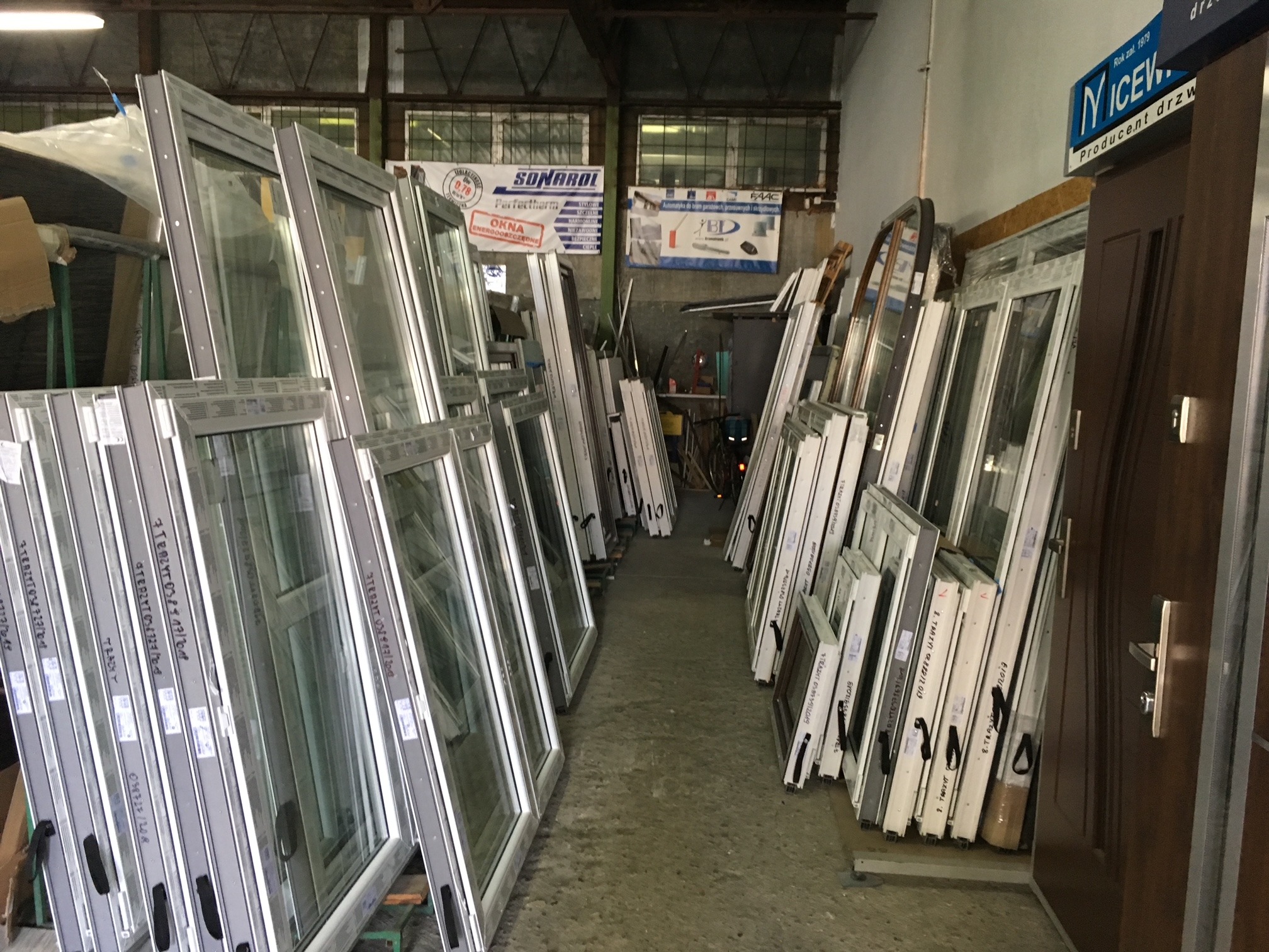 Magazyn z dużą ilością nowych okien i drzwi z ramami w kolorze szarym, opartych o ściany i pod ścianami, z widocznymi etykietami i oznaczeniami na ramach.