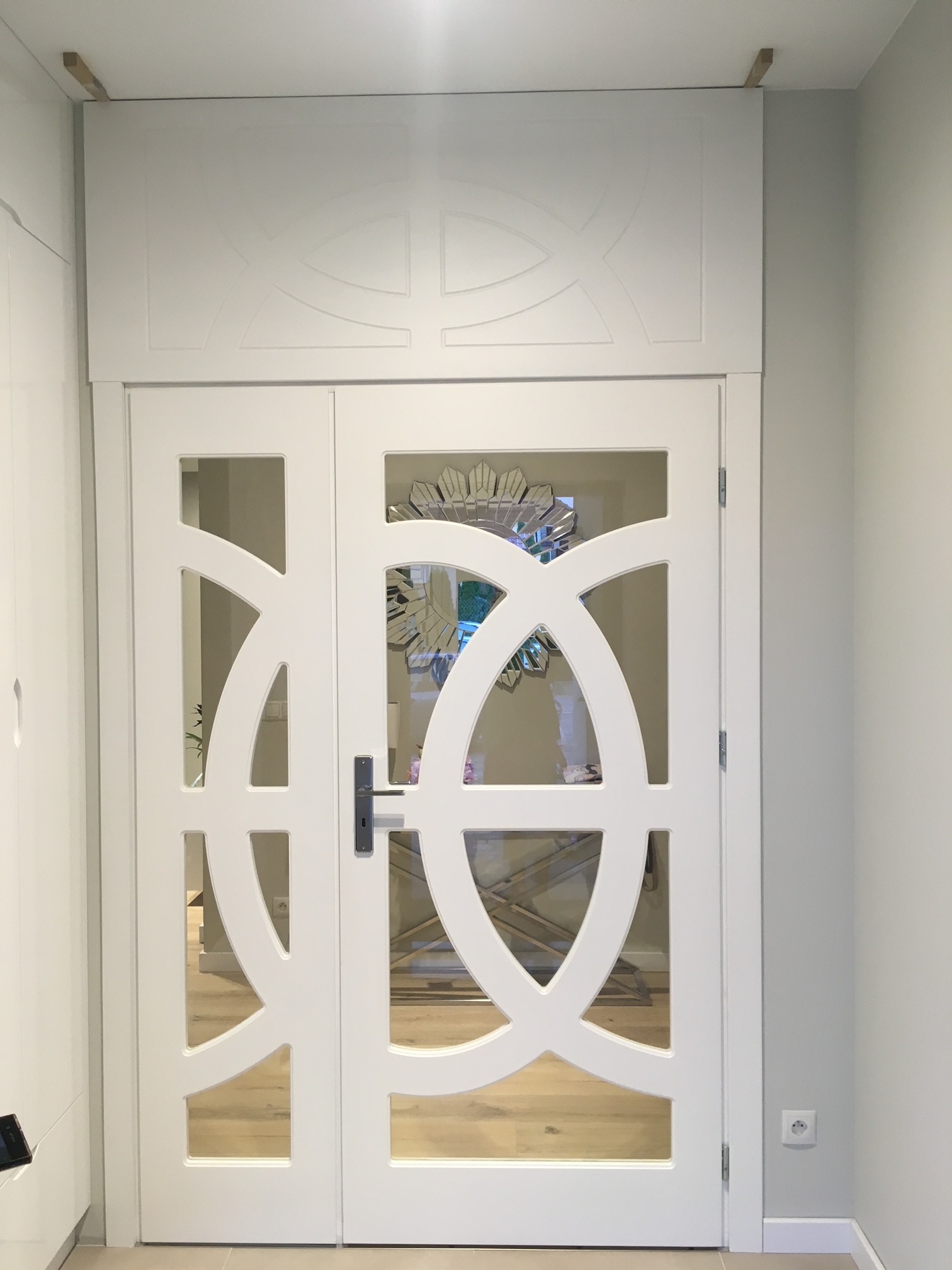 Białe dwuskrzydłowe drzwi wewnętrzne z przeszklonymi dekoracyjnymi elementami, zamontowane w jasnym pomieszczeniu z drewnianą podłogą.