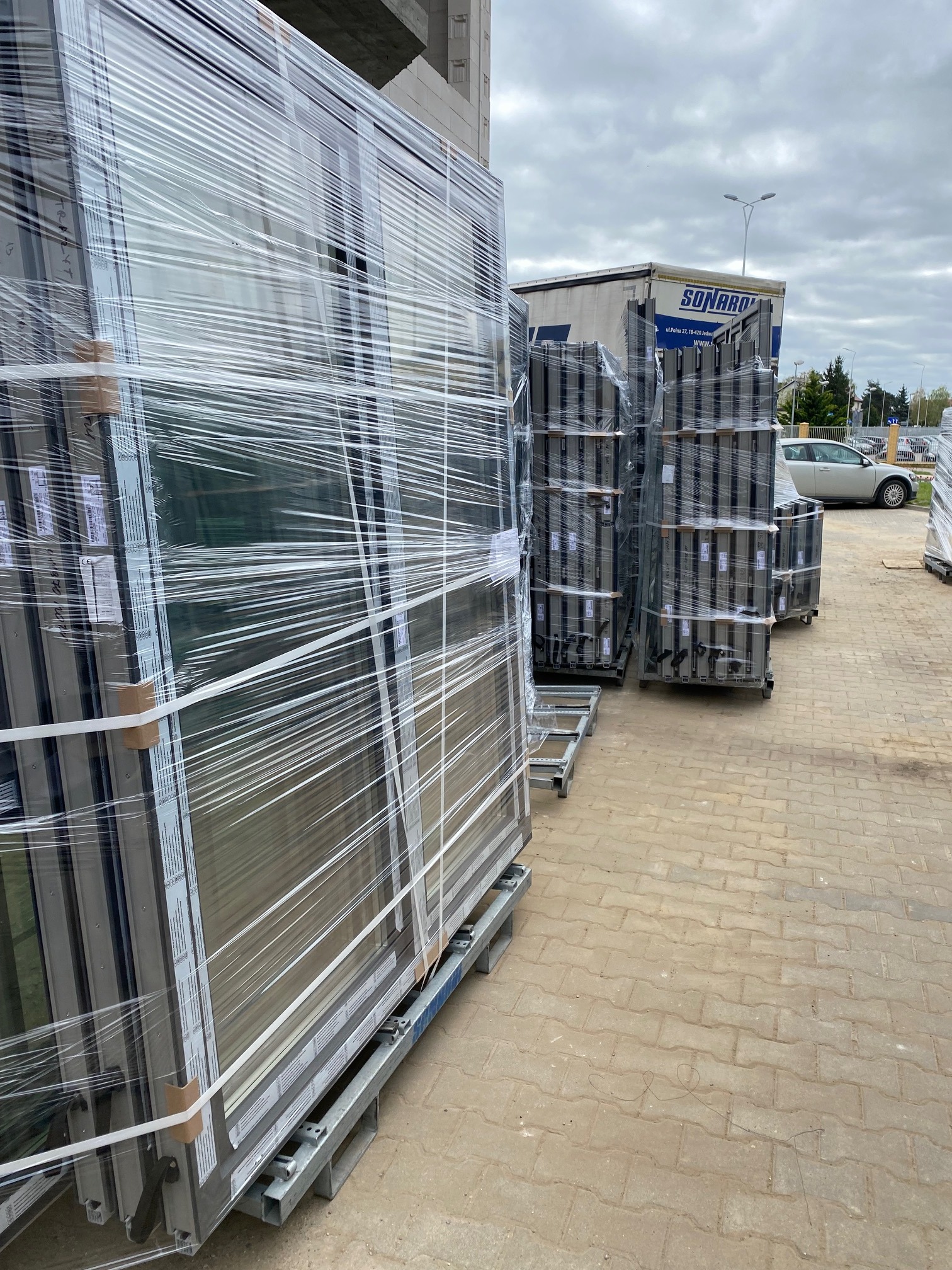 Pakiety zabezpieczonych folią okien i ram okiennych na paletach, przygotowane do transportu na placu budowy, z widoczną ciężarówką w tle.