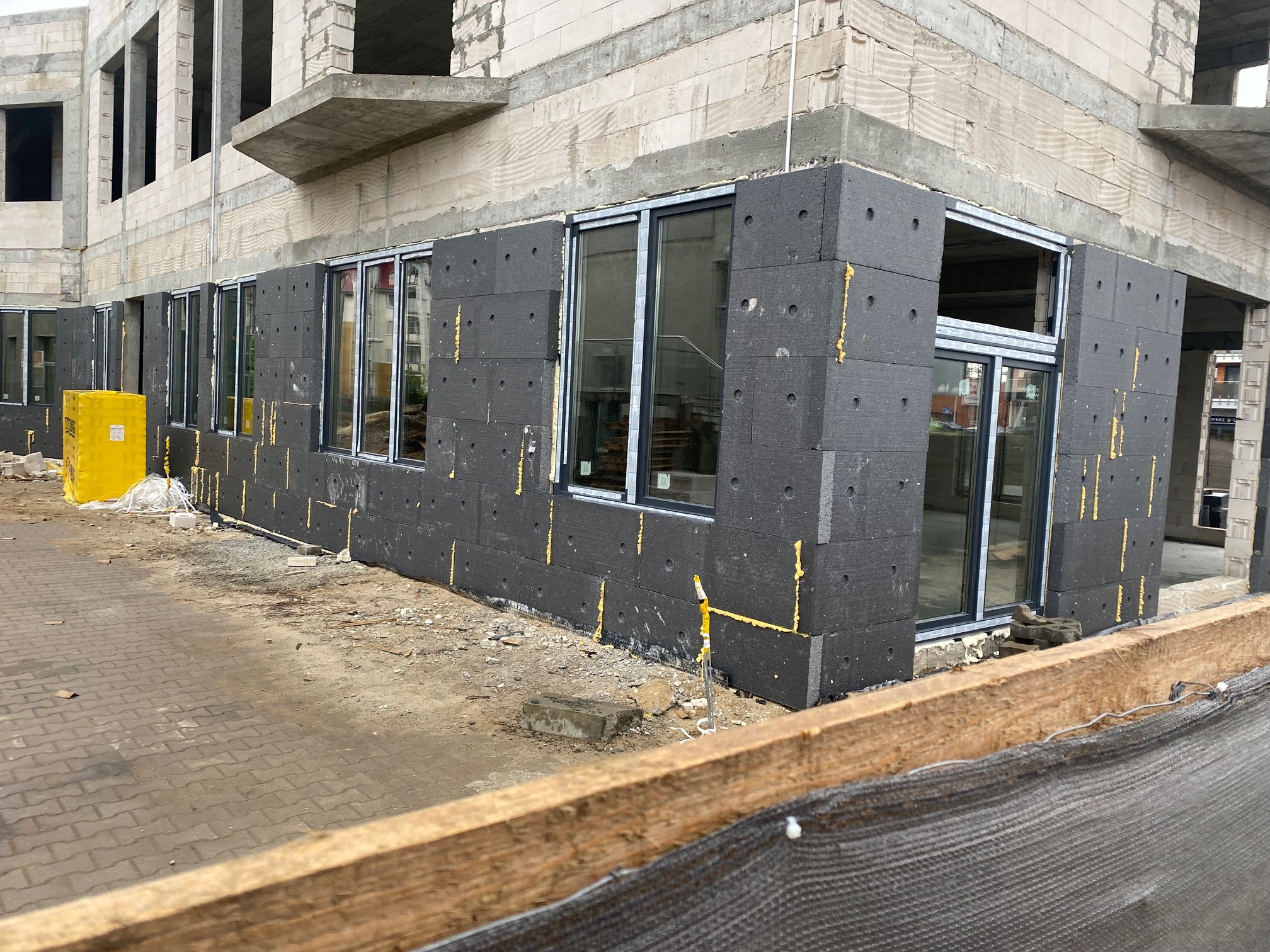 Montaż okien w surowym budynku z widocznym ociepleniem styropianem grafitowym i żółtą pianką montażową, widok z zewnątrz na plac budowy.