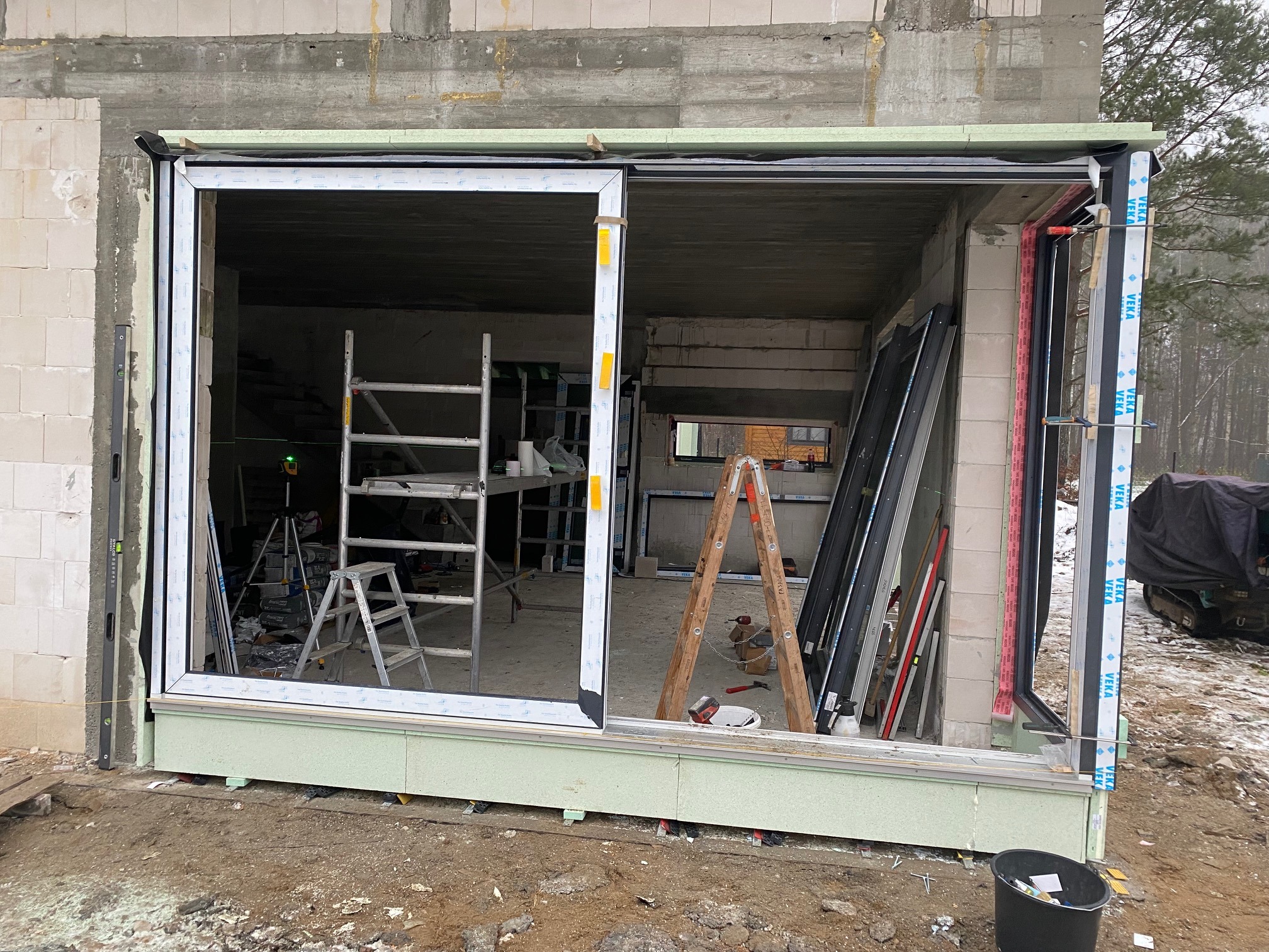 Montaż dużego okna przesuwnego w surowym stanie budynku, widoczne rusztowania, poziomica, narzędzia i elementy konstrukcyjne.