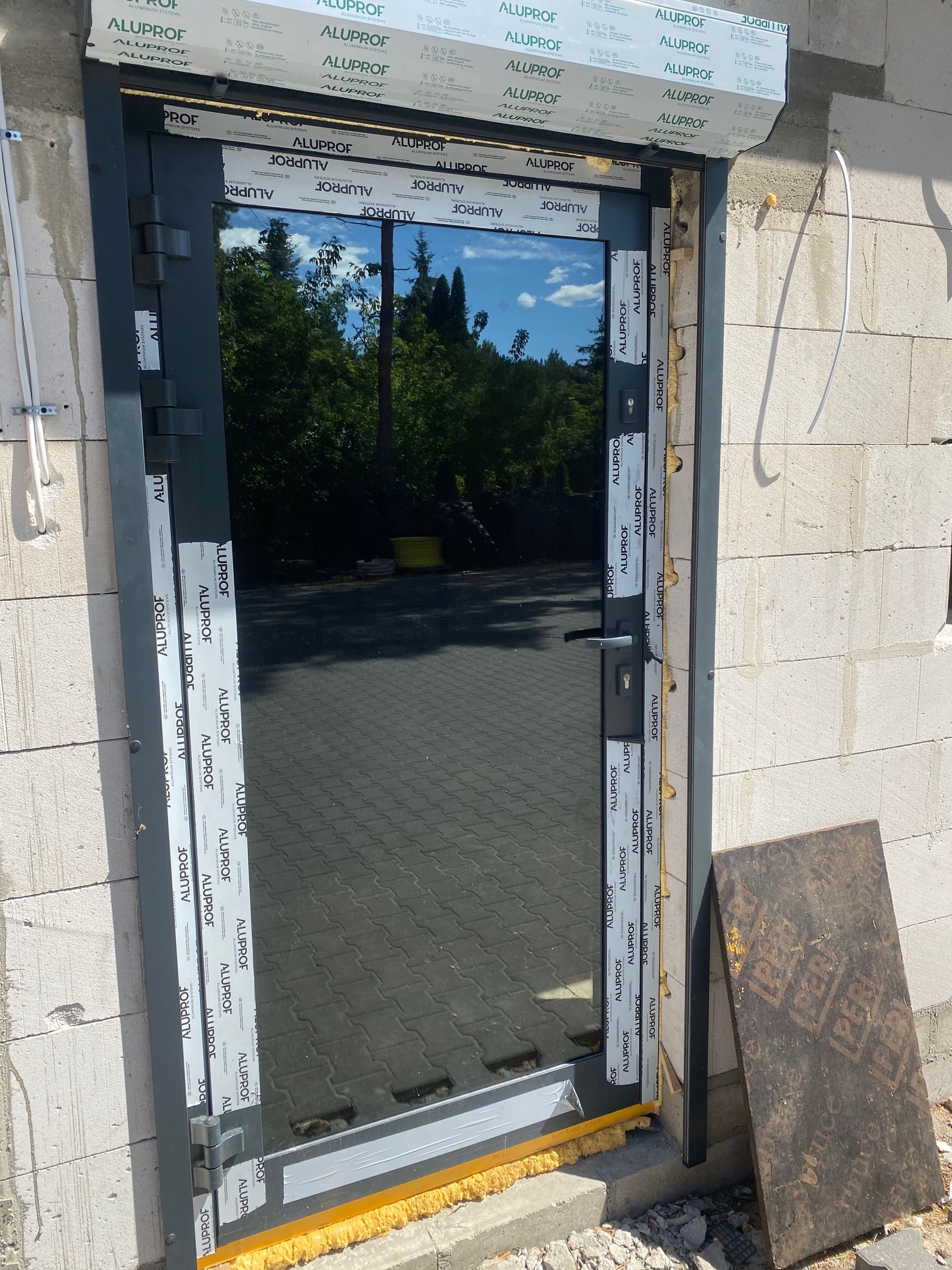 Montaż zewnętrznych drzwi stalowych w surowym stanie, widoczne pianka montażowa, bloczki betonowe i odblask w szybie.