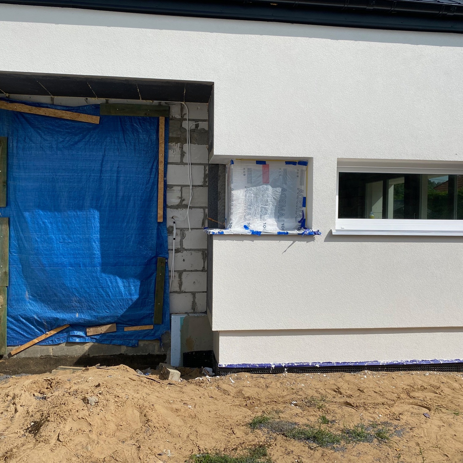 Budynek w trakcie budowy: zabezpieczony otwór okienny niebieską plandeką, okno zafoliowane, obok widoczne kolejne okno, piasek na pierwszym planie.