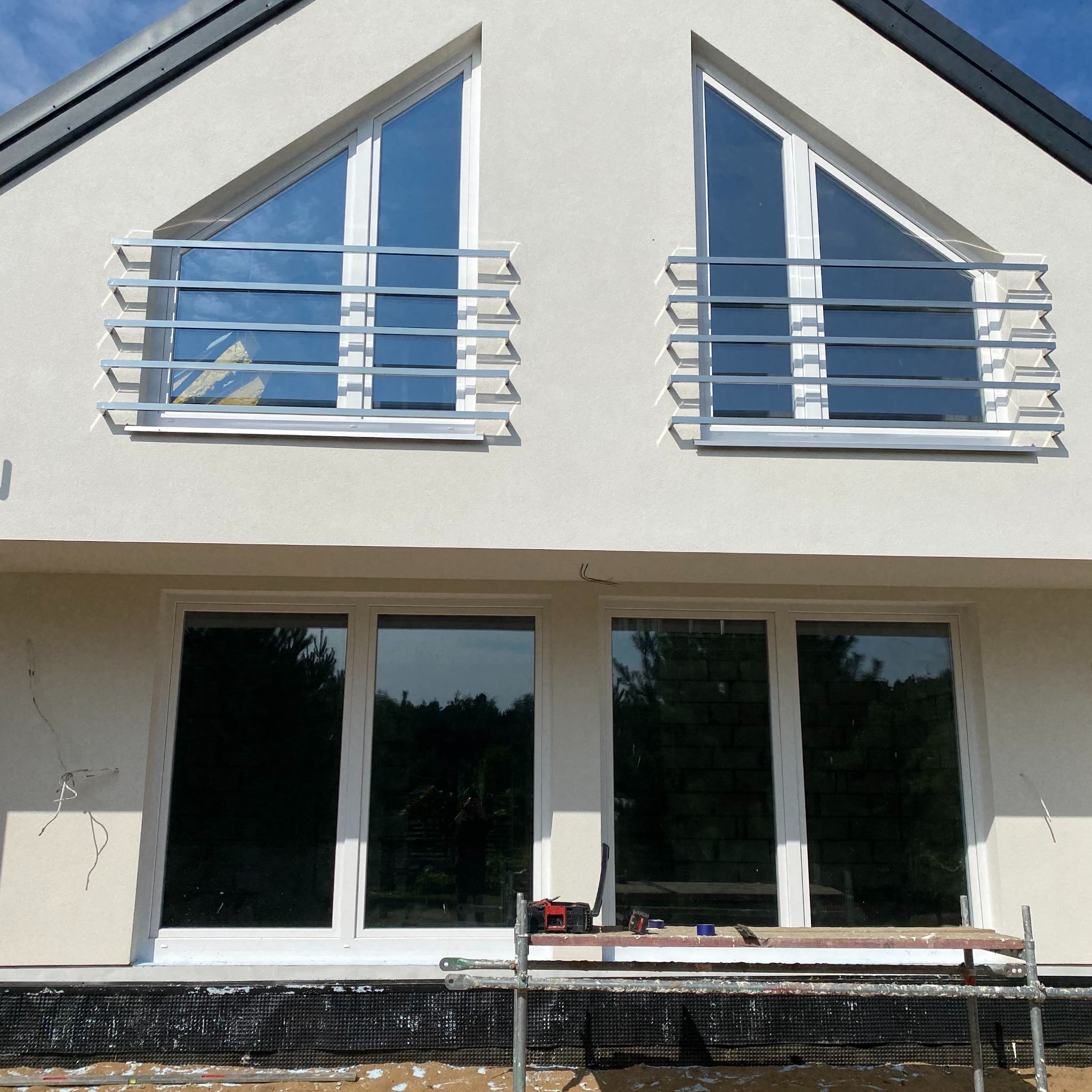 Elewacja domu z dwoma trójkątnymi oknami na górze i dwoma prostokątnymi poniżej, zabezpieczonymi metalowymi balustradami, widoczne rusztowanie i narzędzia sugerujące prace wykończeniowe.