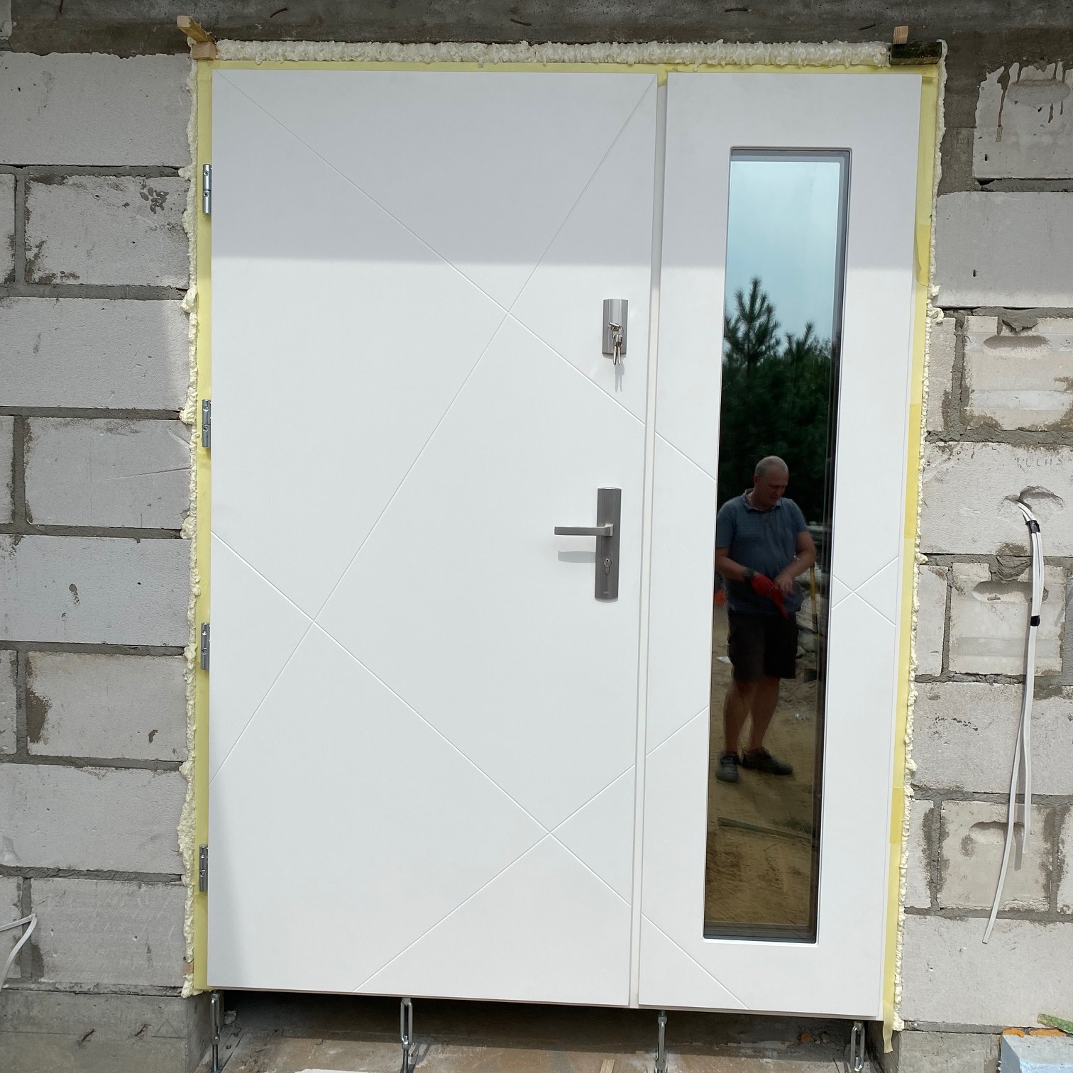 Białe drzwi zewnętrzne z szybą, zamontowane w surowym murze, uszczelnione pianką montażową, widoczne klucze w zamku i odbicie osoby w szybie.