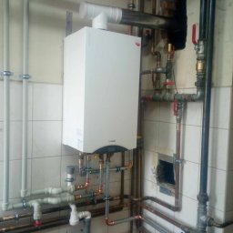 INSTAL-SANIT Adam Zaręba - Nowy, biały piec gazowy Termet zamontowany na ścianie z białych kafelków, z widocznymi miedzianymi i szarymi rurami instalacji, oraz fragmentem sylwetki instalatora w czerwonej koszulce w dolnej...