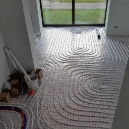 Białe rury ogrzewania podłogowego ułożone spiralnie na folii z czerwoną siatką w nowym budynku z widokiem na zewnątrz przez duże okno, obok ściany leżą narzędzia i puszki.