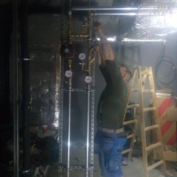 INSTAL-SANIT Adam Zaręba - Instalator w trakcie montażu pionowej instalacji z rur miedzianych i stalowych, z manometrami i pompą obiegową, na tle drabiny i izolowanych ścian, w pomieszczeniu o słabym oświetleniu.