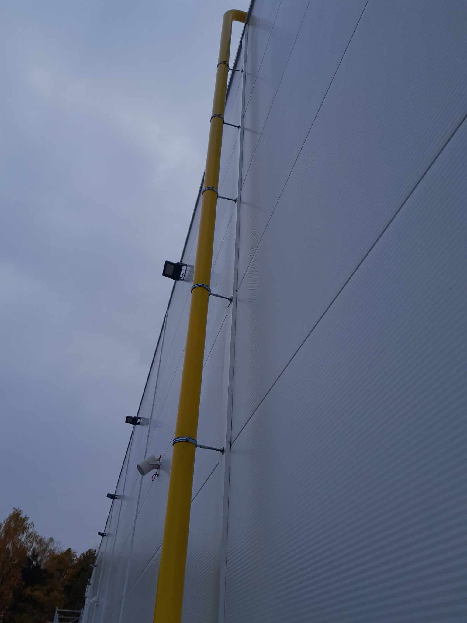 Żółta rura spustowa przymocowana do ściany hali przemysłowej z widocznymi lampami zewnętrznymi i pochmurnym niebem w tle.