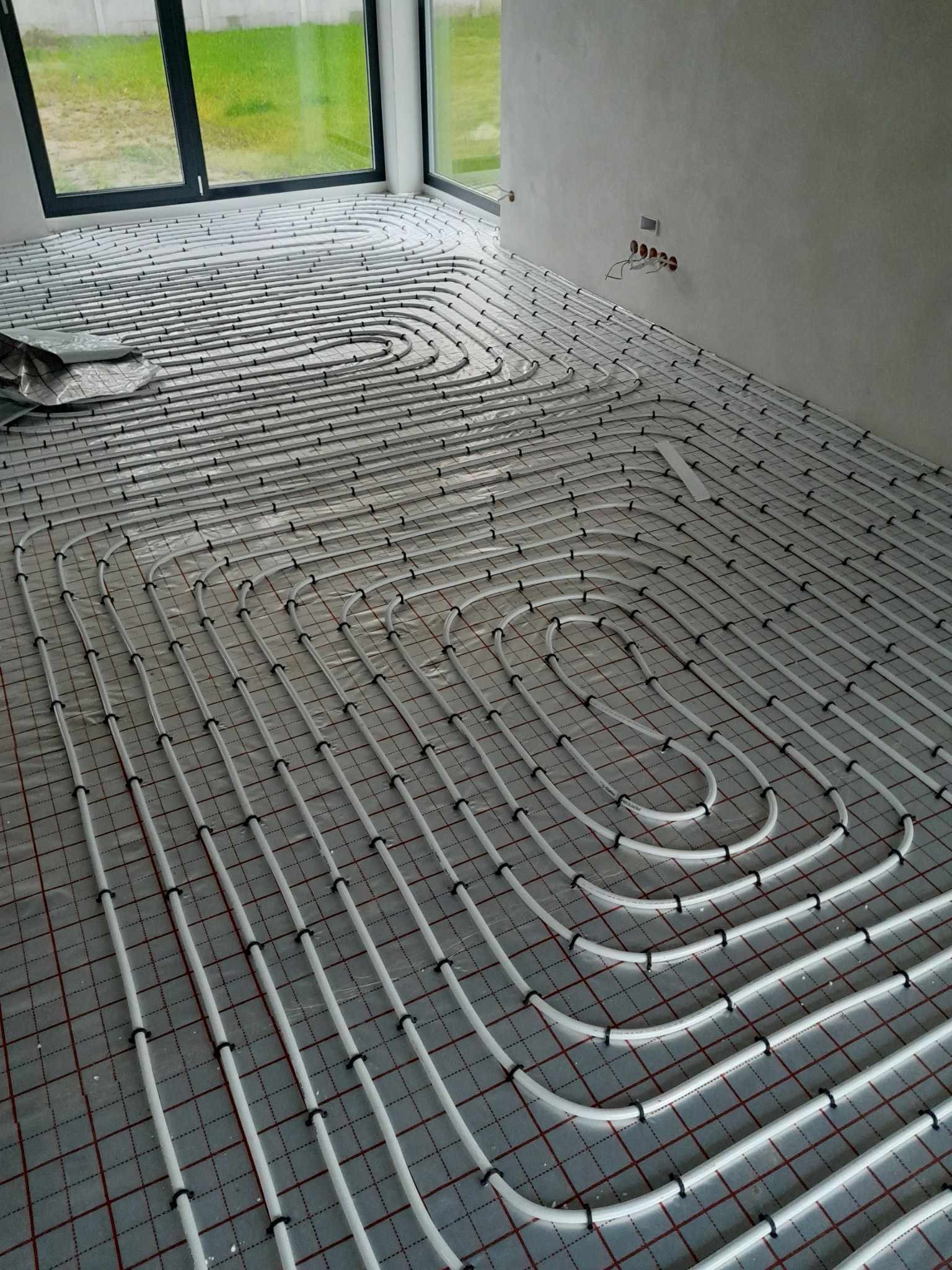 Białe rury ogrzewania podłogowego ułożone w pętle na folii z czerwoną siatką w surowym wnętrzu z dużym oknem