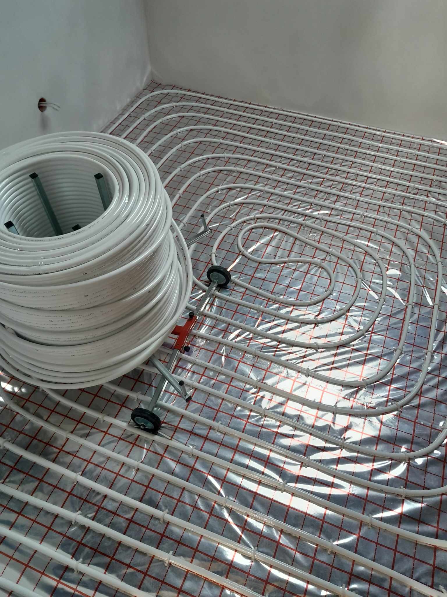 Białe rury ogrzewania podłogowego rozłożone na folii z siatką, zwój rur na wózku obok, surowy stan wykończenia wnętrza.