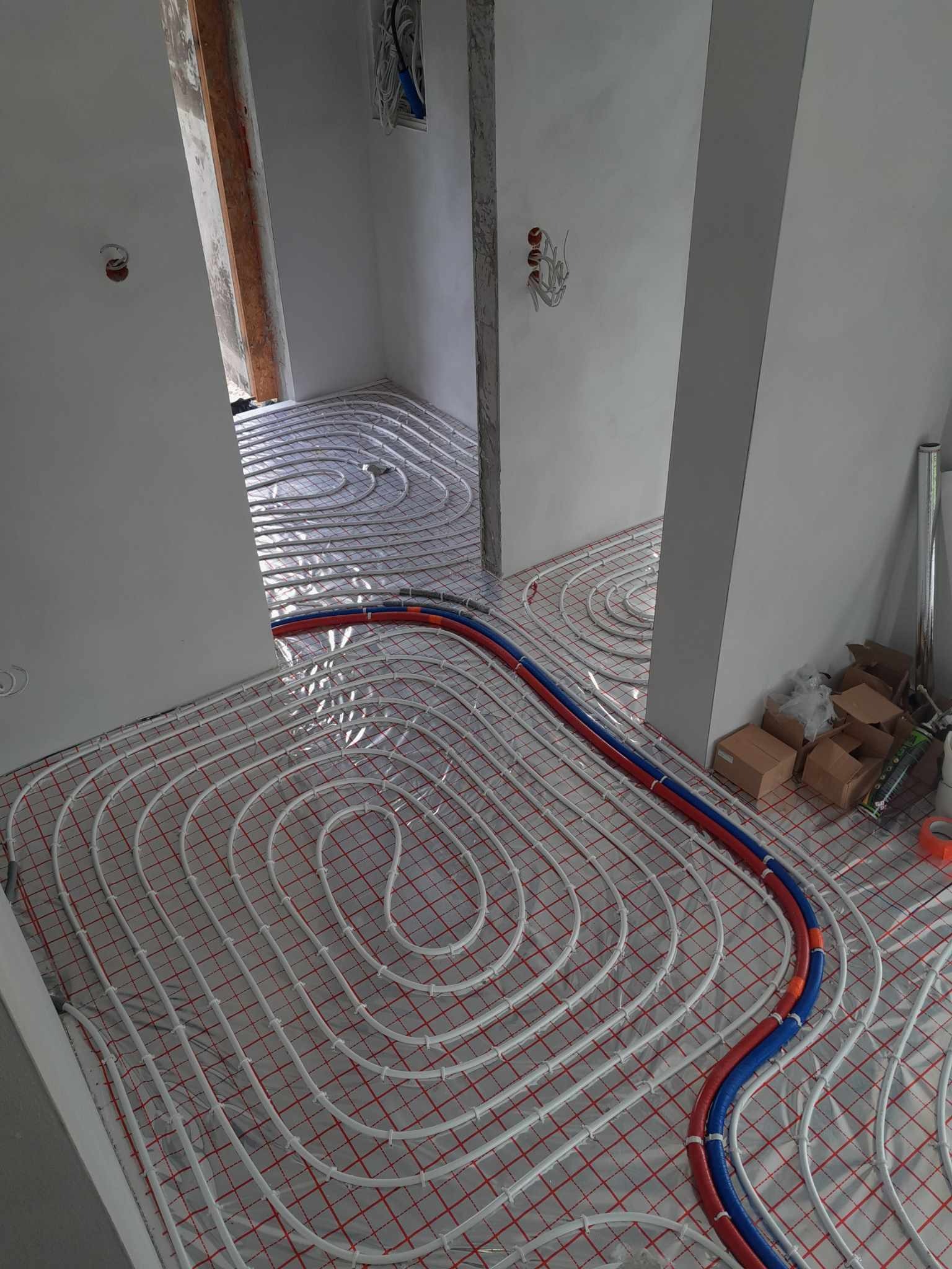 Białe rurki systemu grzewczego podłogowego ułożone na folii z czerwoną siatką, widoczne rury doprowadzające w kolorze niebieskim i czerwonym, surowy stan wykończenia wnętrza.