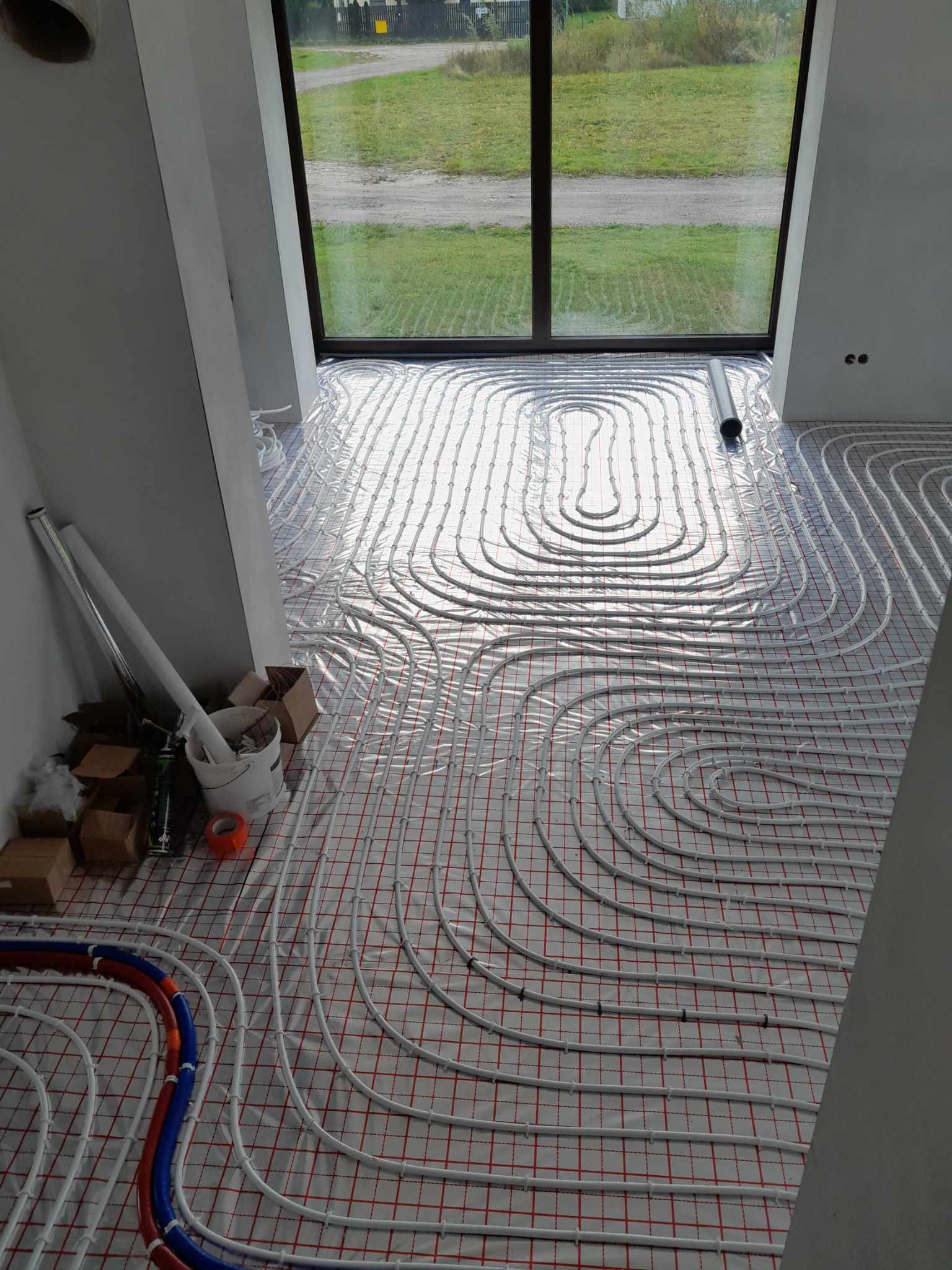Białe rury ogrzewania podłogowego ułożone spiralnie na folii z czerwoną siatką w nowym budynku z widokiem na zewnątrz przez duże okno, obok ściany leżą narzędzia i puszki.