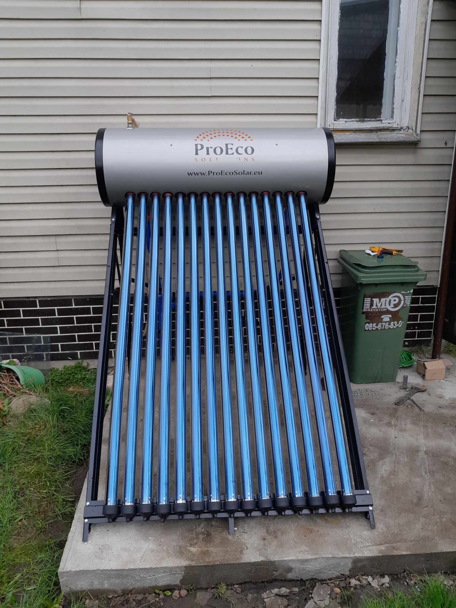 Kolektor słoneczny rurowy z srebrnym zbiornikiem i niebieskimi rurami próżniowymi, zamontowany na betonowej podstawie przed budynkiem z sidingiem; obok stoi zielony kosz na śmieci.