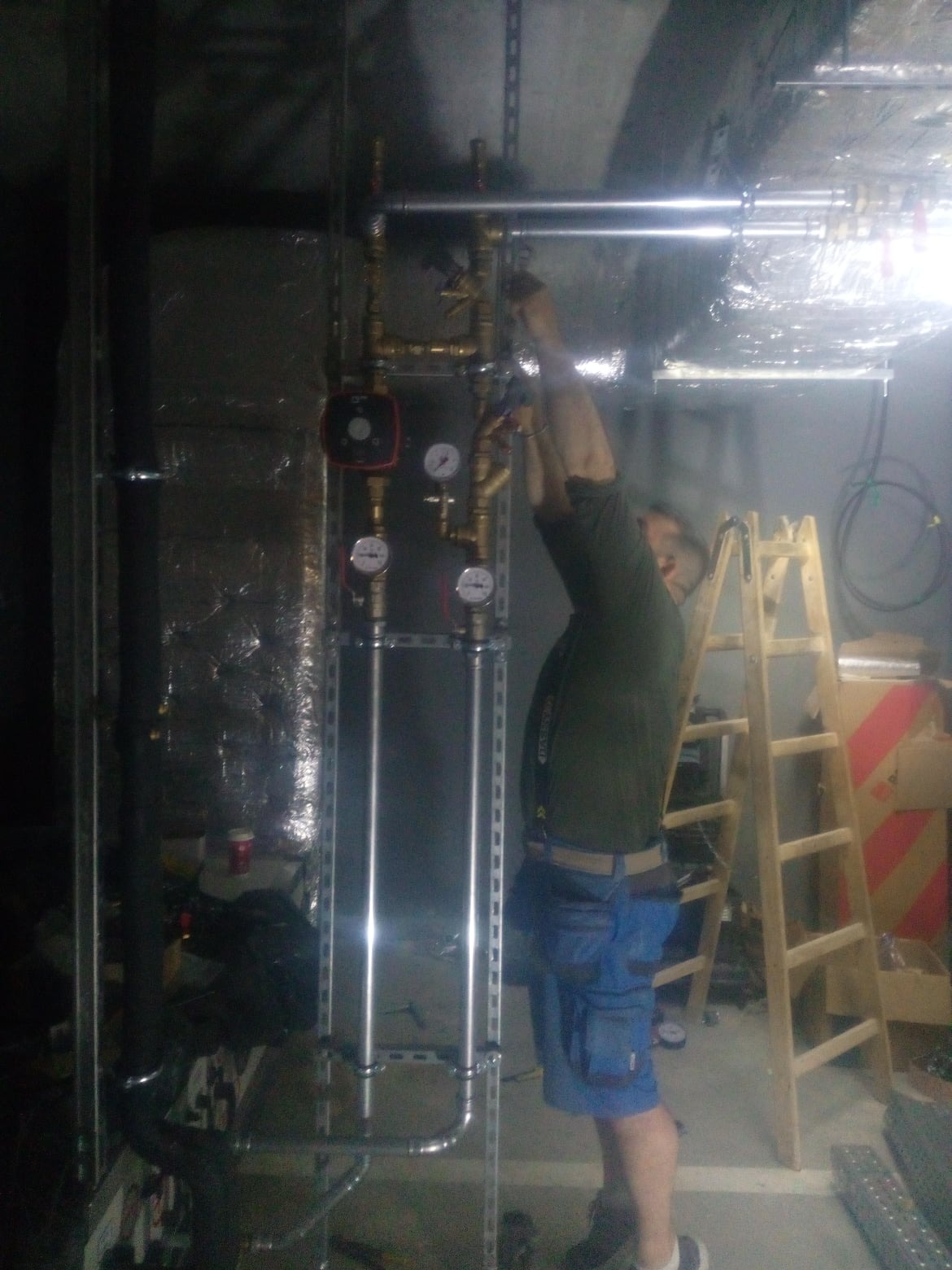 Instalator w trakcie montażu pionowej instalacji z rur miedzianych i stalowych, z manometrami i pompą obiegową, na tle drabiny i izolowanych ścian, w pomieszczeniu o słabym oświetleniu.