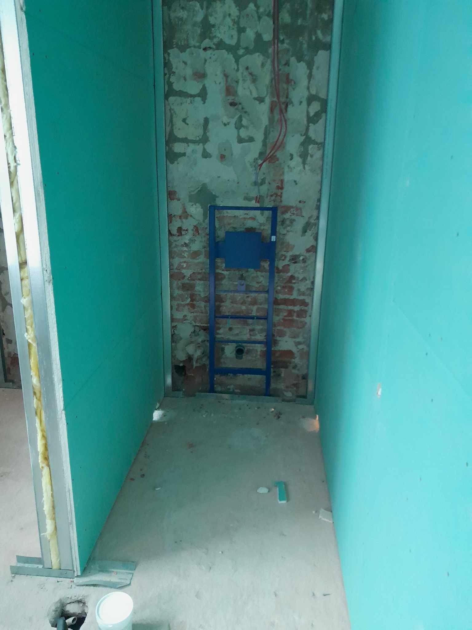 Wnętrze w trakcie remontu łazienki: widoczne płyty gipsowo-kartonowe w kolorze niebieskim, metalowy stelaż podtynkowy do WC na tle starej, odsłoniętej cegły, surowa podłoga betonowa, puszka z farbą...