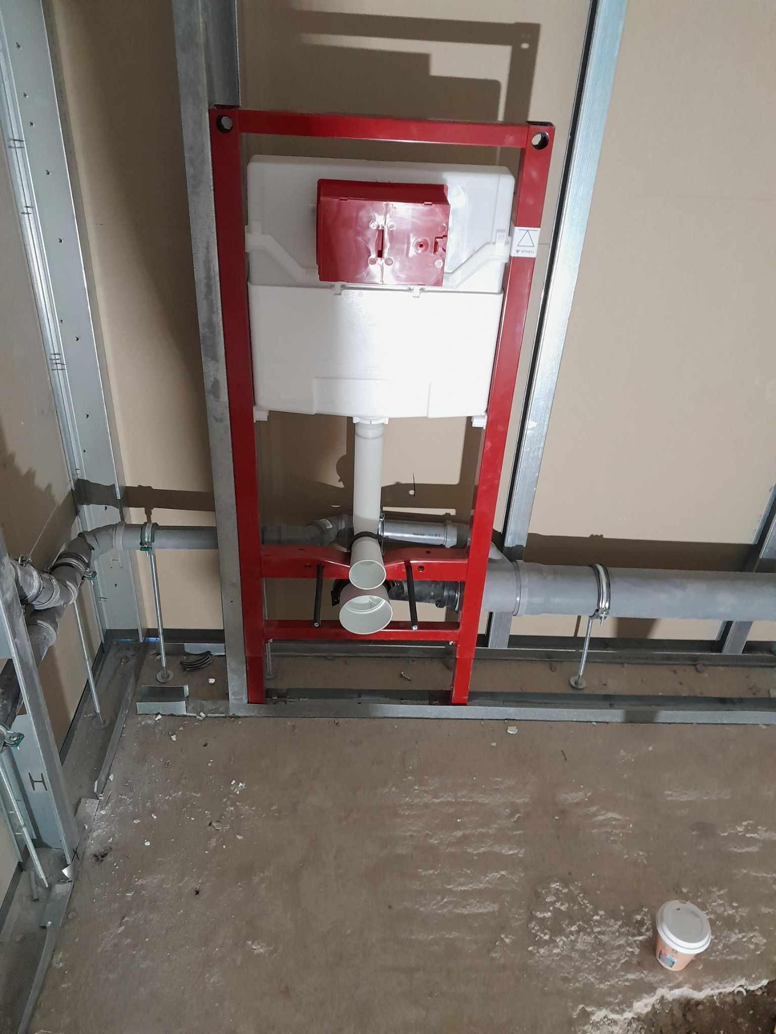 Stelaż podtynkowy WC z czerwoną ramą, widoczne rury kanalizacyjne i elementy konstrukcji ściany działowej w surowym stanie.