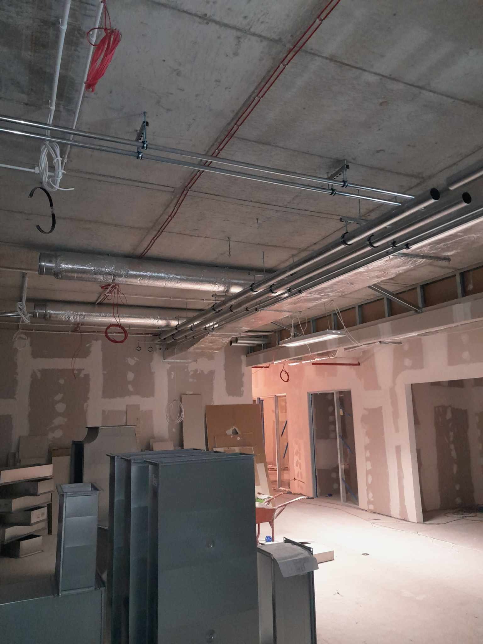 Wnętrze w stanie surowym z widocznymi elementami instalacji wentylacyjnej i elektrycznej pod sufitem, metalowe rury i kanały wentylacyjne, czerwone i białe przewody, ściany przygotowane do malowania.