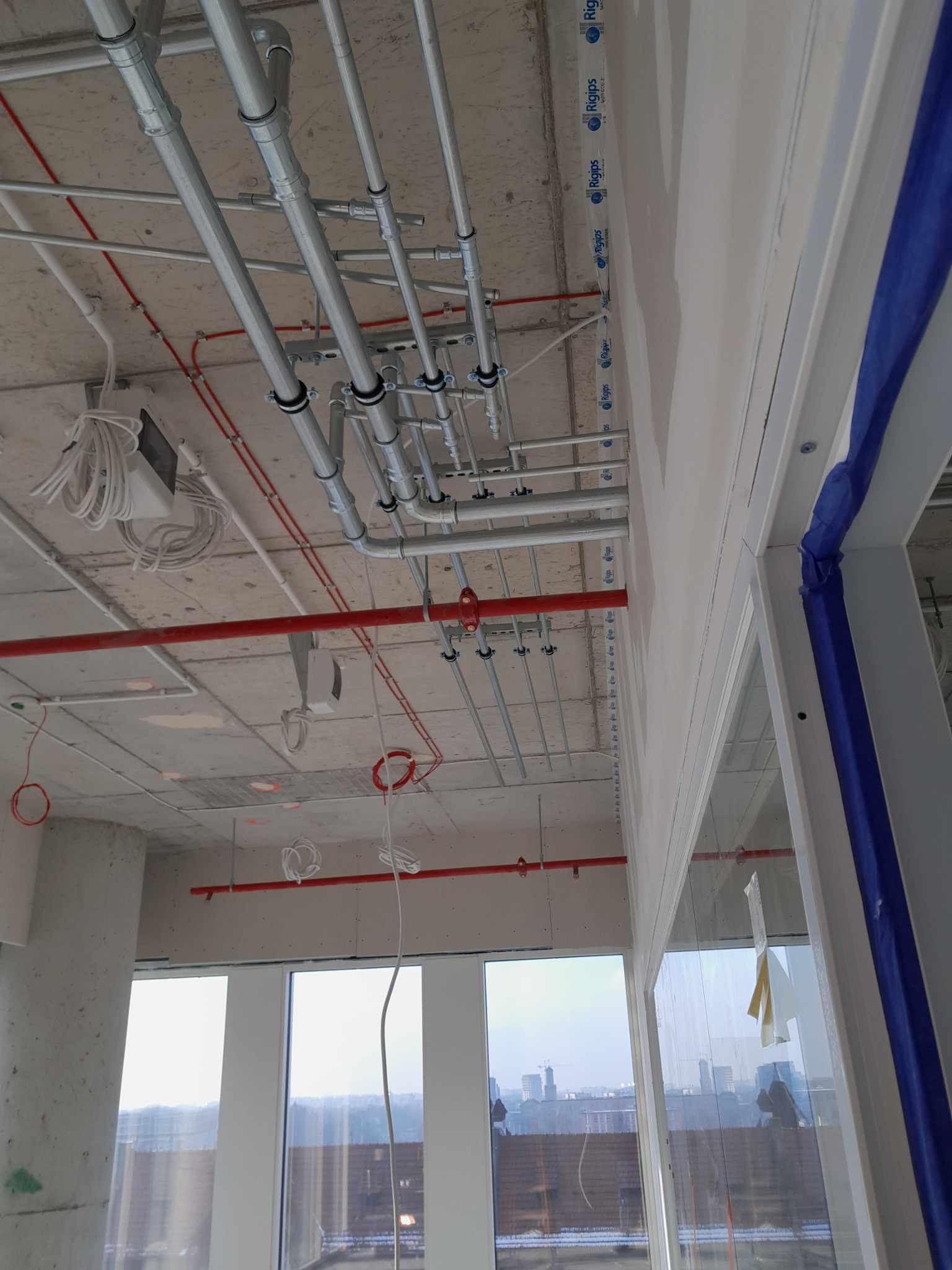 Widok na sufit w trakcie prac wykończeniowych, z widocznymi rurami instalacyjnymi (metalowe, szare i czerwone) oraz przewodami elektrycznymi, na tle surowego betonu i okien z widokiem na miasto.