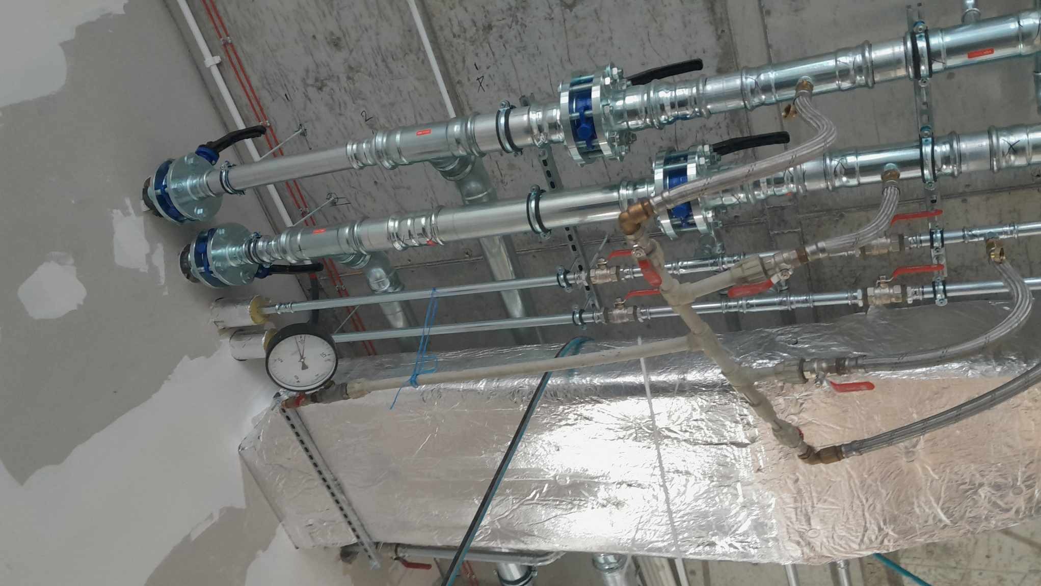 Widok z dołu na sufit z zamontowanymi rurami, zaworami i manometrem, instalacja w surowym stanie, widoczne łączenia i mocowania.