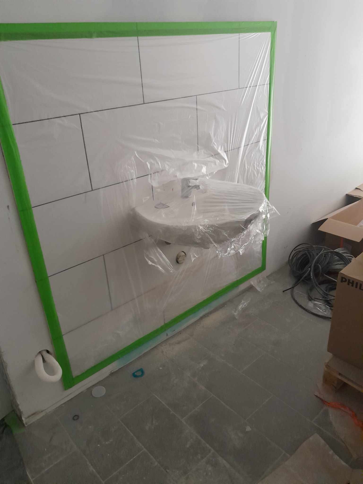 Zabezpieczona folią i zieloną taśmą umywalka z baterią na tle białych płytek podczas remontu łazienki, widoczne przewody i kartony.