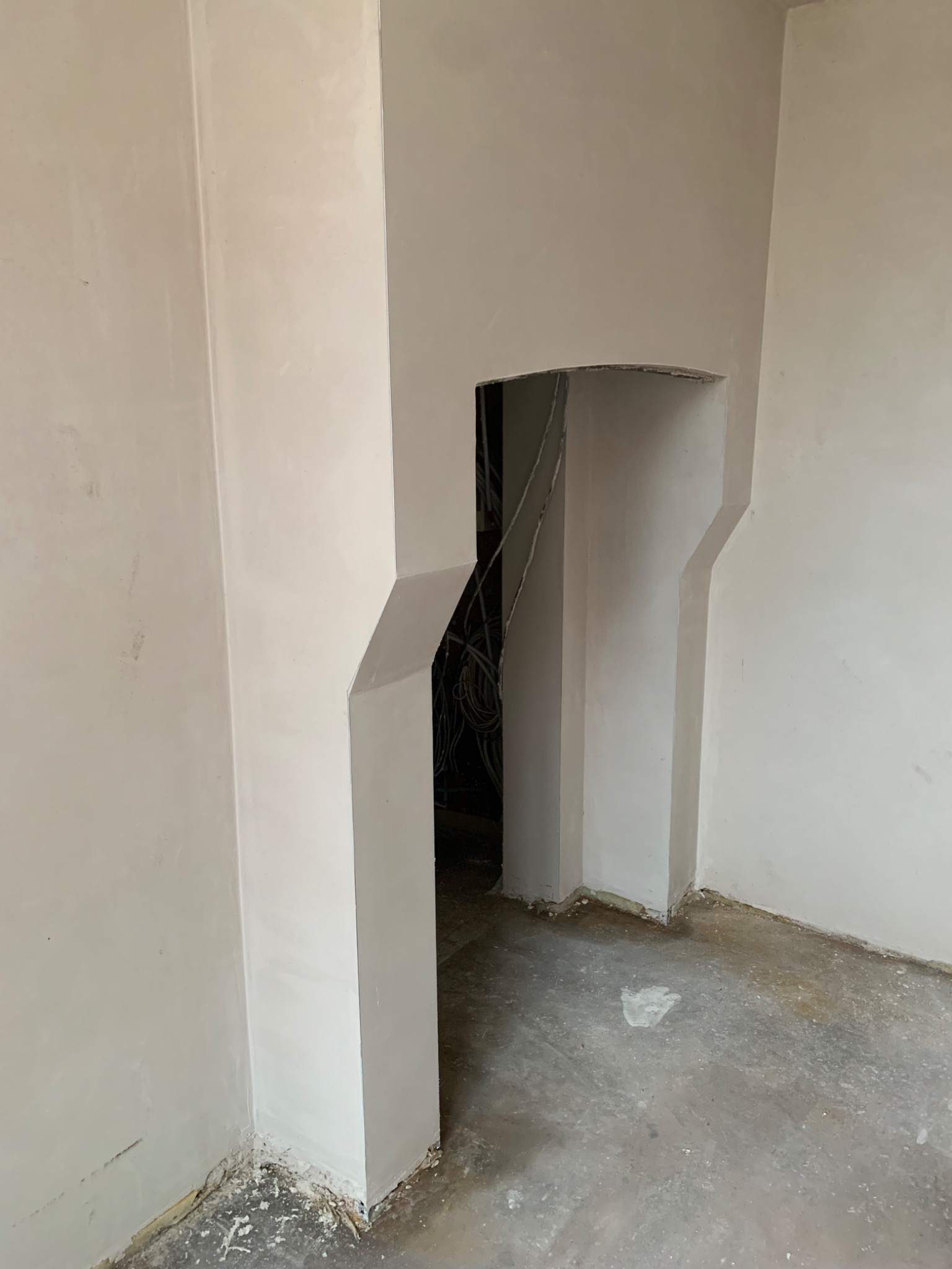 Wnętrze w trakcie prac wykończeniowych: świeżo otynkowane ściany w jasnym odcieniu, widoczne kable w otworze ściany, surowa podłoga. Charakterystyczne załamania ścian.