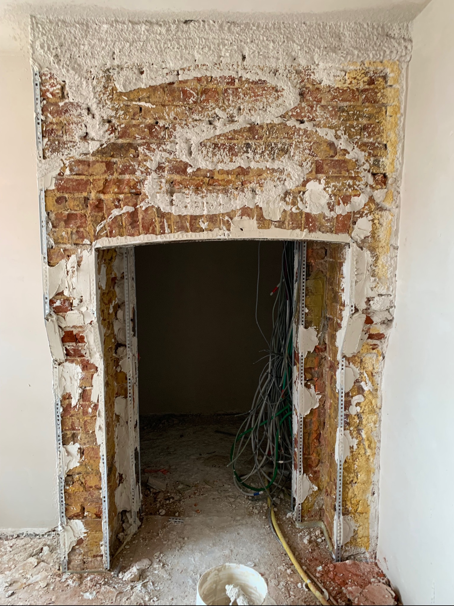 Ościeżnica z cegły w trakcie szpachlowania, widoczne metalowe profile i przewody elektryczne. Fragment ściany z nałożoną warstwą gładzi. Prace wykończeniowe wewnątrz budynku.