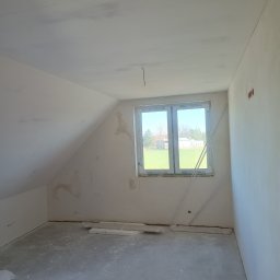 MAXBUD - Pokój na poddaszu po szpachlowaniu, ściany przygotowane do malowania. Widoczne okno z widokiem na zewnątrz i fragment instalacji elektrycznej.