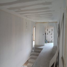 MAXBUD - Korytarz w trakcie prac wykończeniowych: ściany i sufit po szpachlowaniu, przygotowane do malowania, widoczne płyty gipsowe i schody betonowe. Surowy stan.