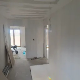 Wnętrze w trakcie prac wykończeniowych: surowy sufit po szpachlowaniu, ściany przygotowane do malowania, widoczny sprzęt malarski i płyty gipsowe.