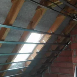 Montaż metalowego stelażu pod okładzinę na poddaszu, widoczne drewniane krokwie, folia paroizolacyjna i fragment komina z cegły.